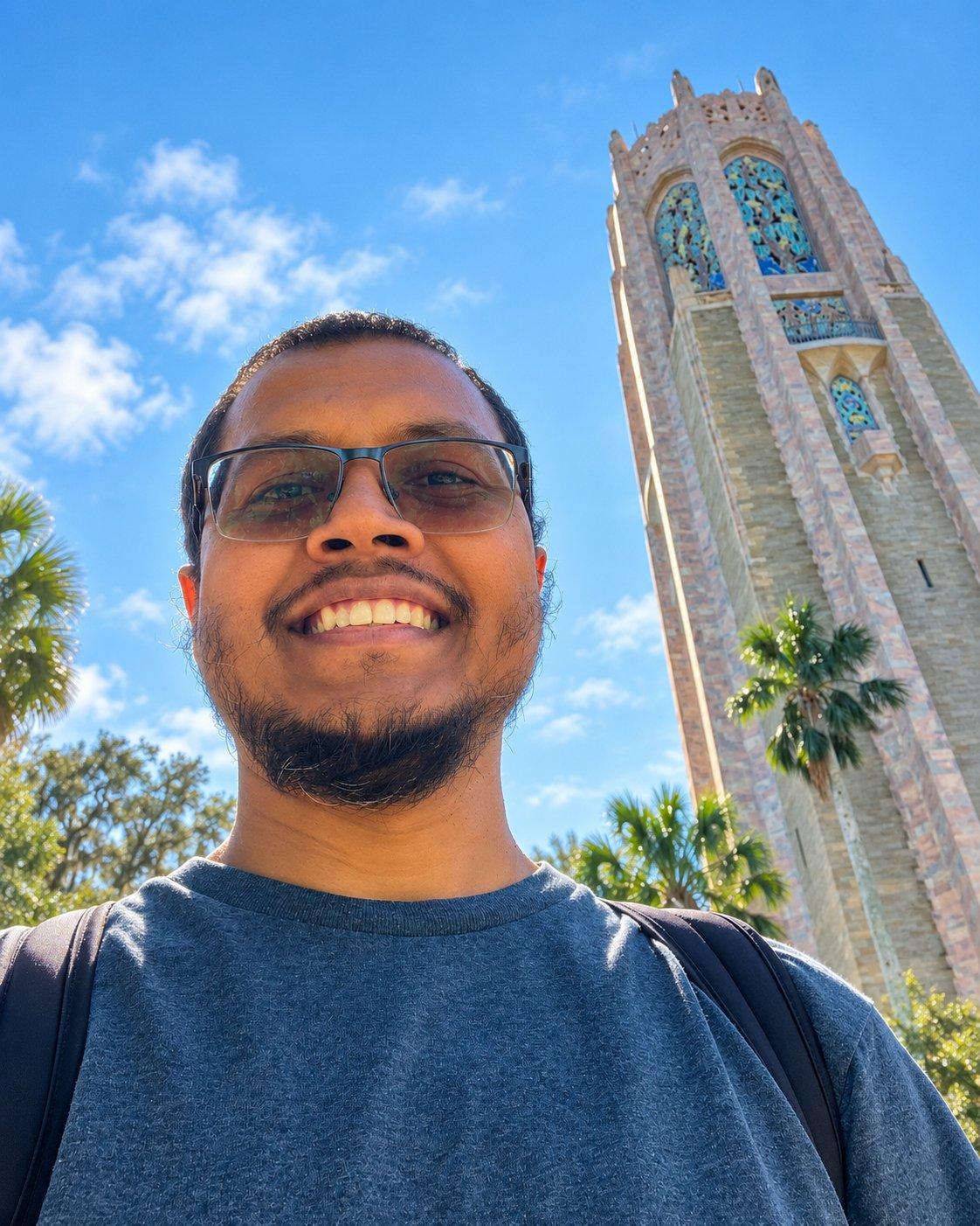 Ash smiling outdoors in front of a tall stone tower on a sunny day